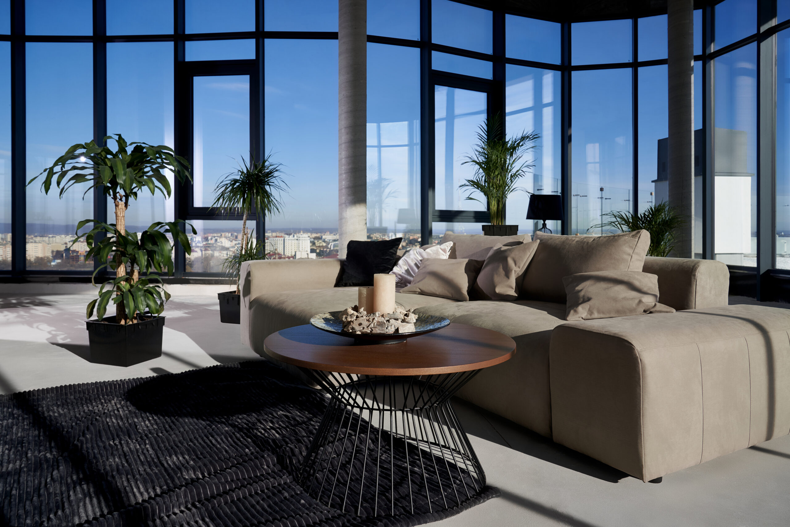 Front view of fashionable interior spacious room with large panoramic window with incredible view on city and cozy grey sofa with pillows and green flowerpots. Concept of modern interior.